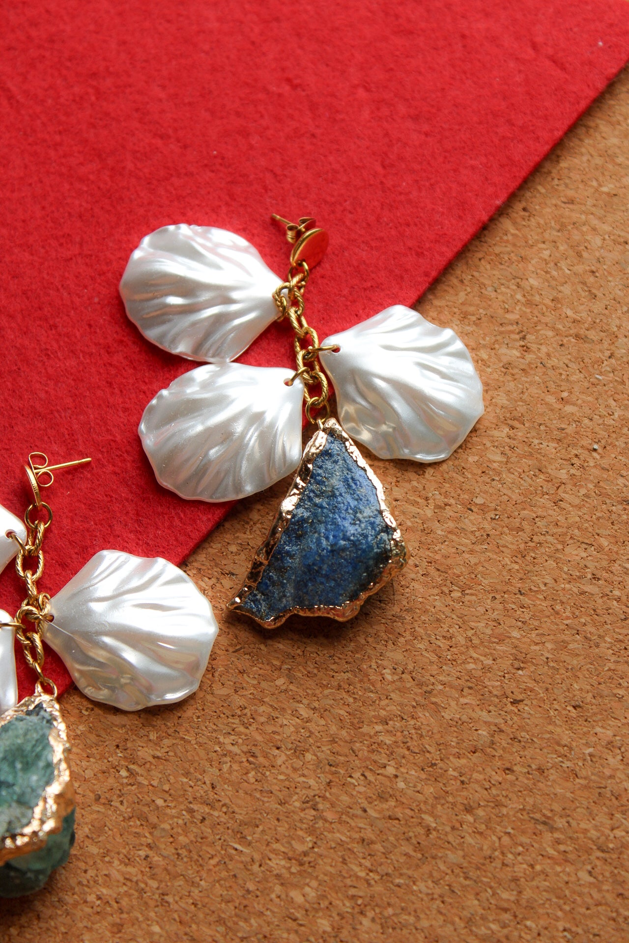 A pair of handmade dangle earrings featuring white mother-of-pearl flower petals, gold-tone metal wire wrapping, and raw stone drops. One stone is a textured blue agate, and the other is a greenish-blue geode slice, displayed on a split red and brown background.
Pantallas artesanales de Marveyra hechas a mano en Puerto Rico. Diseños playeros, únicos y personalizables con elementos removibles que permiten crear diferentes combinaciones o utilizar los colgantes por separado. Joyería diferente