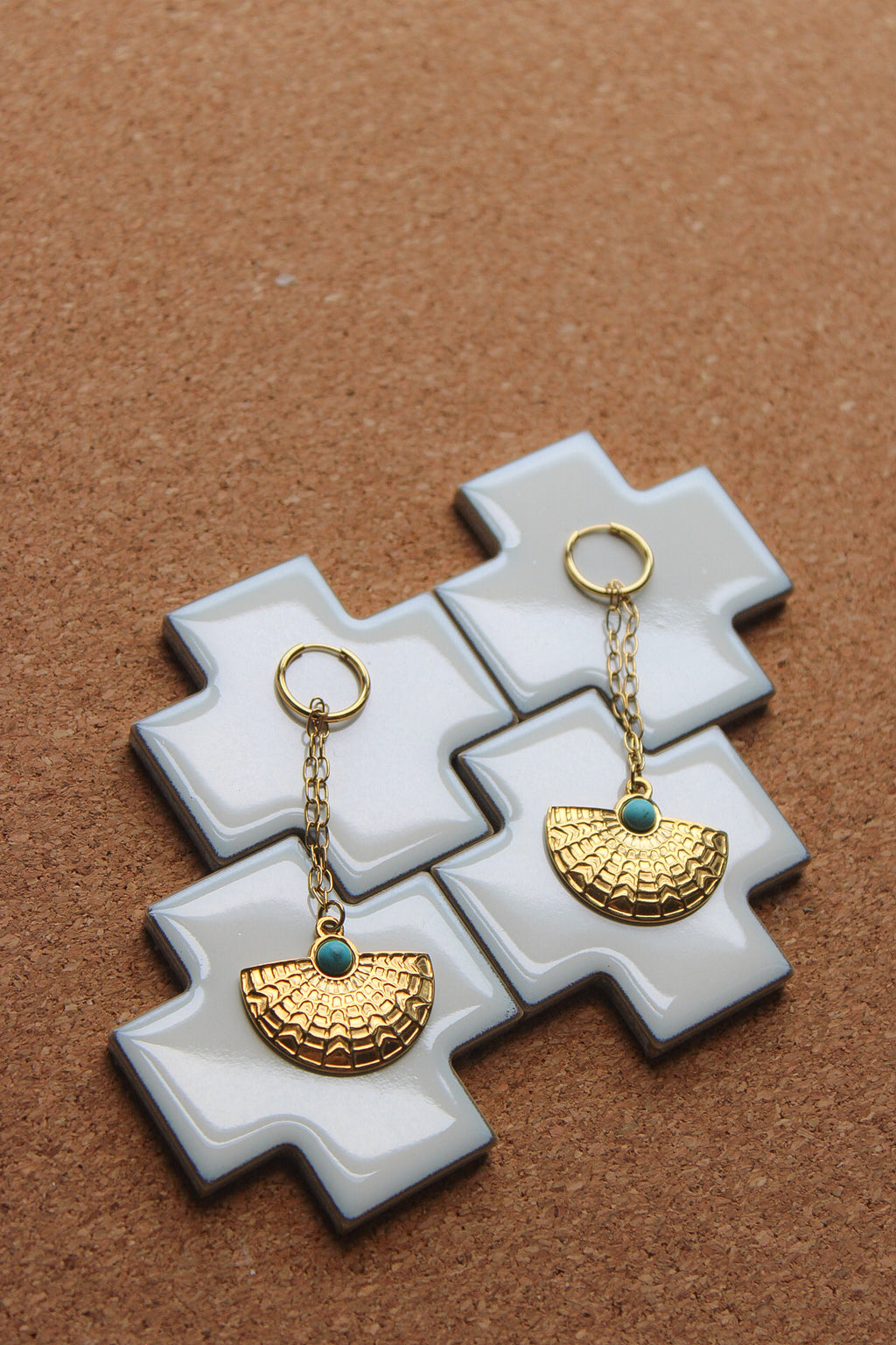 A pair of gold-tone dangle earrings with textured semicircle charms and small turquoise blue beads, displayed on white cross-shaped tiles over a brown cork background.Pantallas artesanales de Marveyra, hechas a mano en Puerto Rico con materiales resistentes. Este diseño de joyería alternativa y unisex cuenta con elementos removibles que permiten personalizar la pieza o utilizar los colgantes por separado como collar. Accesorios originales creados para quienes buscan destacar con un estilo diferente.