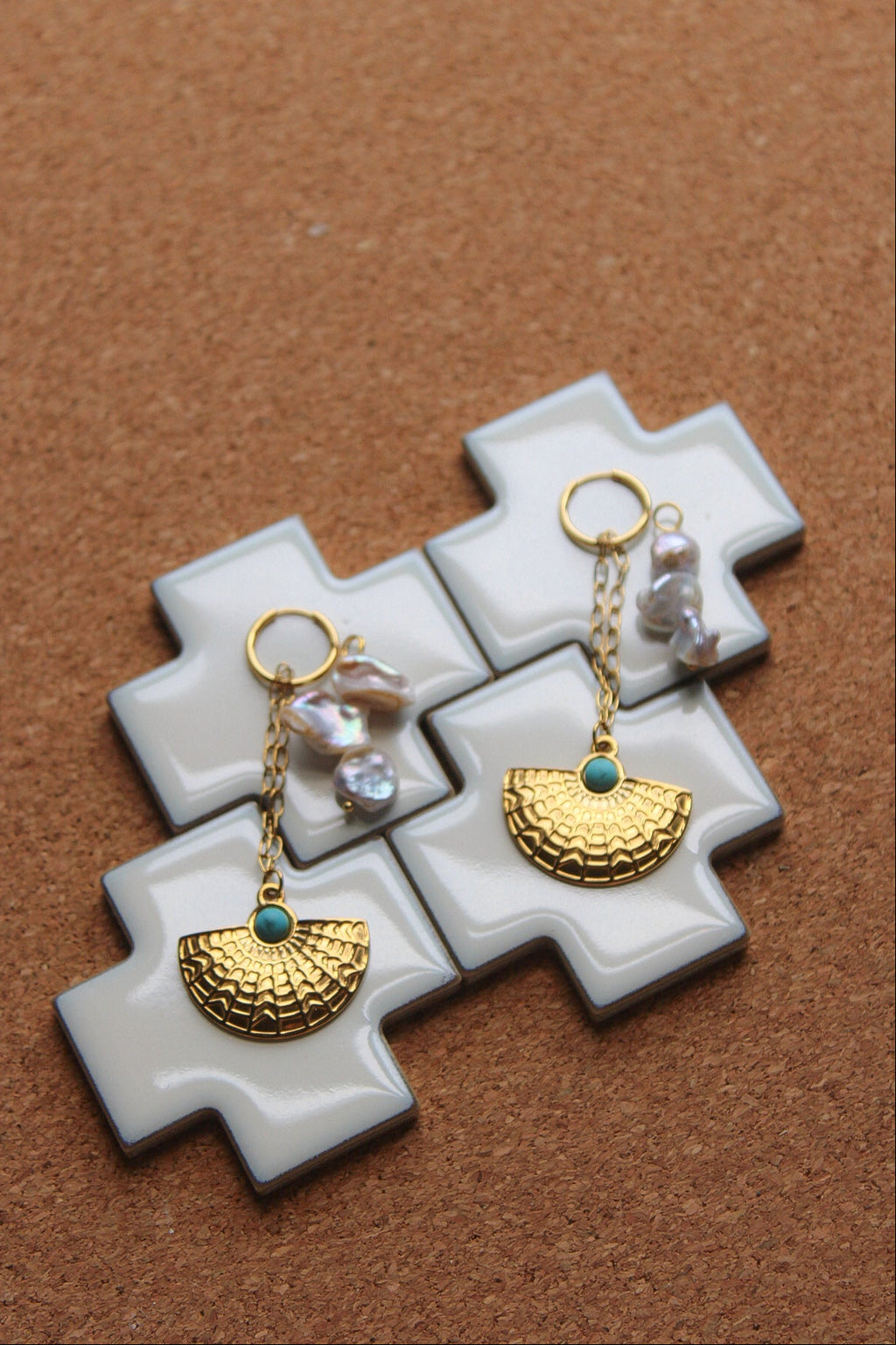A pair of gold-toned dangle earrings with textured semicircle charms, small turquoise blue stones, and irregular white pearls, displayed on a white cross-shaped stand over a brown cork background.
Pantallas artesanales de Marveyra, hechas a mano en Puerto Rico con materiales resistentes. Este diseño de joyería alternativa y unisex cuenta con elementos removibles que permiten personalizar la pieza o utilizar los colgantes por separado como collar. Accesorios originales creados para quienes buscan destacar co