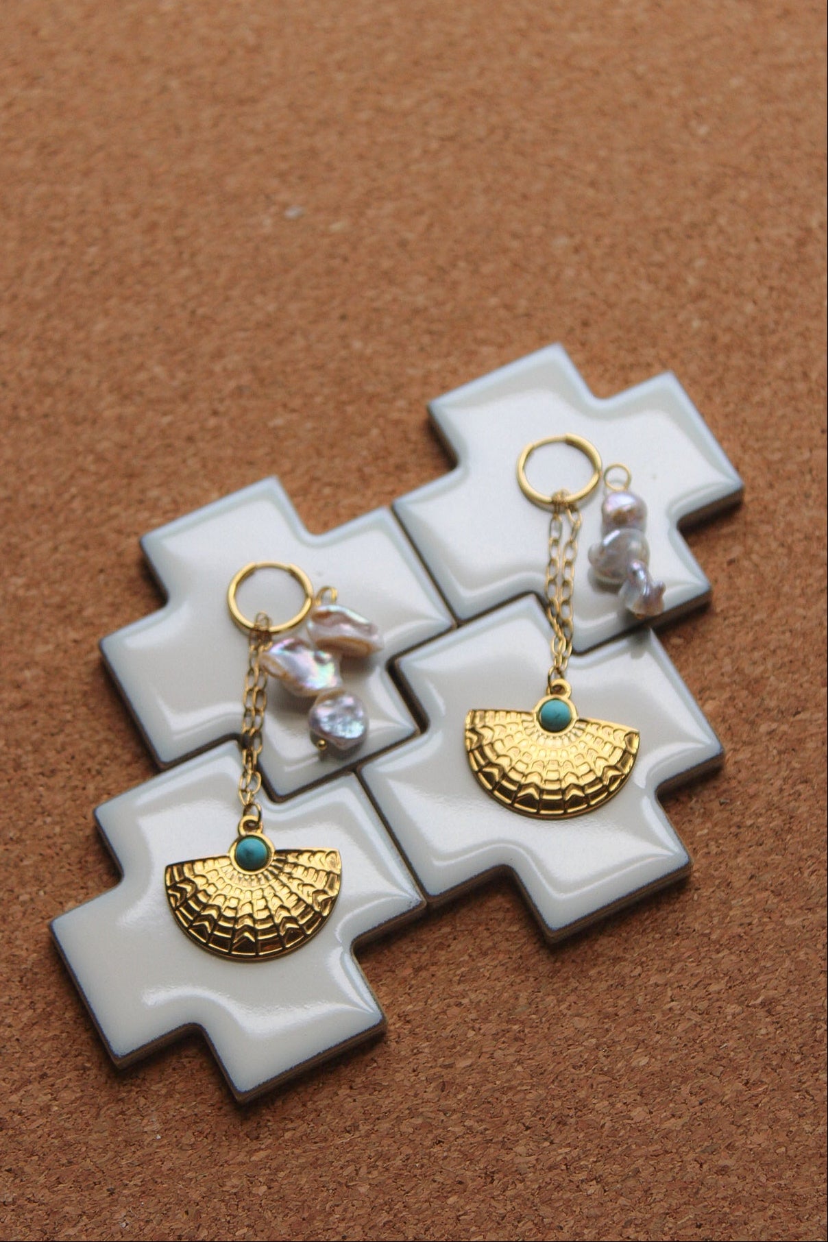 A pair of gold-toned dangle earrings with textured semicircle charms, small turquoise blue stones, and irregular white pearls, displayed on a white cross-shaped stand over a brown cork background.
Pantallas artesanales de Marveyra, hechas a mano en Puerto Rico con materiales resistentes. Este diseño de joyería alternativa y unisex cuenta con elementos removibles que permiten personalizar la pieza o utilizar los colgantes por separado como collar. Accesorios originales creados para quienes buscan destacar co