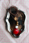 A beaded necklace with a brown cord, gold-toned disc bead, brown wooden beads, a white bead, and a large glossy red bead, displayed on a small silver tray over a pink polka dot fabric background.