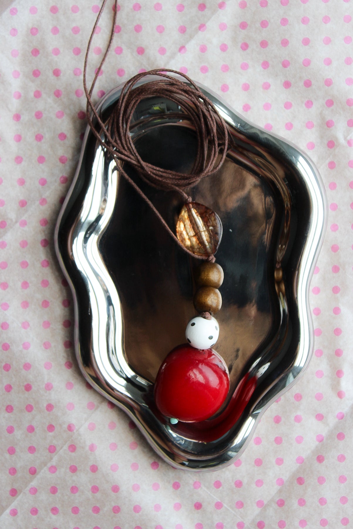 A beaded necklace with a brown cord, gold-toned disc bead, brown wooden beads, a white bead, and a large glossy red bead, displayed on a small silver tray over a pink polka dot fabric background.
