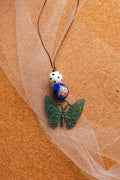 A handmade-style beaded necklace featuring a green filigree butterfly pendant, a blue and white patterned bead, and a white bead with black dots, strung on a brown cord and laid on a cork background with sheer white tulle.