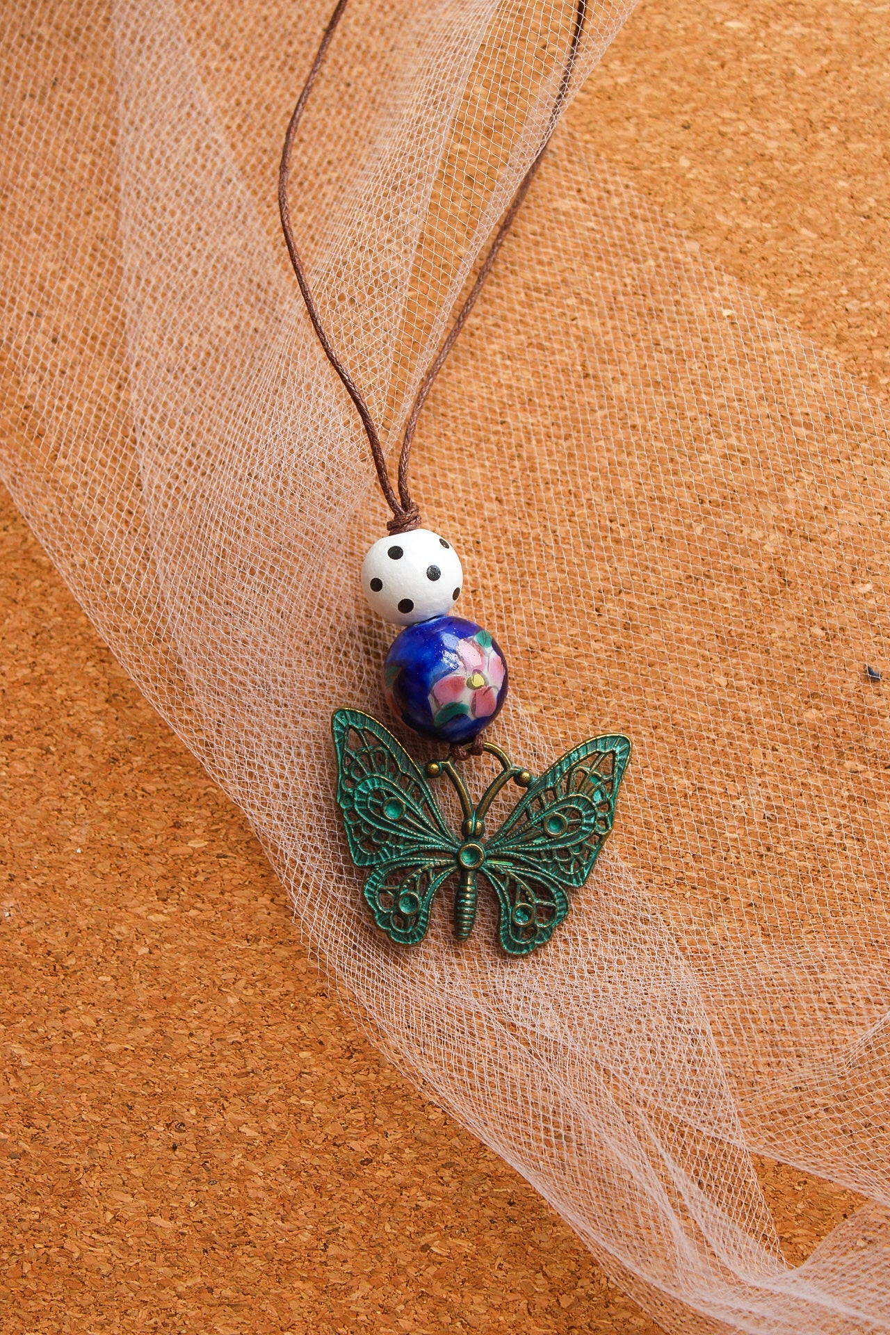 A handmade-style beaded necklace featuring a green filigree butterfly pendant, a blue and white patterned bead, and a white bead with black dots, strung on a brown cord and laid on a cork background with sheer white tulle.