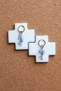 A pair of gold hoop earrings with small light blue flower charms, displayed on white cross-shaped stands against a brown cork background. Pantallas delicadas con gotas de cristales azules cayendo, colgando. Estilo de pantallas sencillas a la moda y diferentes.